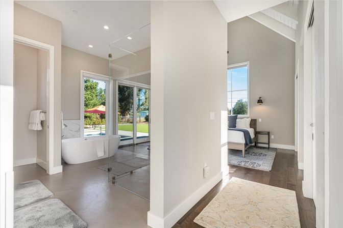 Sunlit modern master suite with open-concept bathroom—freestanding tub and frameless glass shower—sliding glass doors to a green backyard with pool and red umbrella, adjacent vaulted bedroom with hardwood floors and neutral decor.