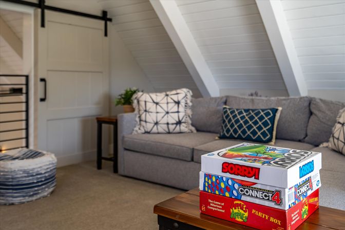 Cozy loft living room with sloped white ceiling, gray sofa with patterned pillows, and a stack of board games on a wooden coffee table ready for game night.