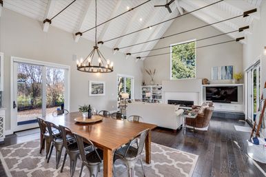 Open-concept dining and living area with vaulted white ceiling and exposed beams, dark hardwood floors, sliding glass doors and tall window to backyard, long wooden dining table with metal chairs under a round chandelier, neutral sofas facing a built-in fireplace and TV — bright, airy space for gatherings.