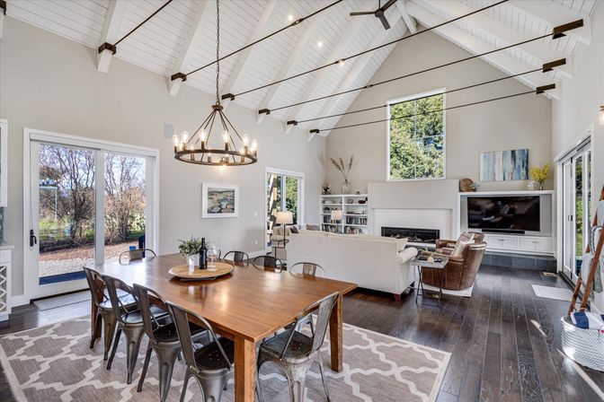 Open-concept dining and living area with vaulted white ceiling and exposed beams, dark hardwood floors, sliding glass doors and tall window to backyard, long wooden dining table with metal chairs under a round chandelier, neutral sofas facing a built-in fireplace and TV — bright, airy space for gatherings.