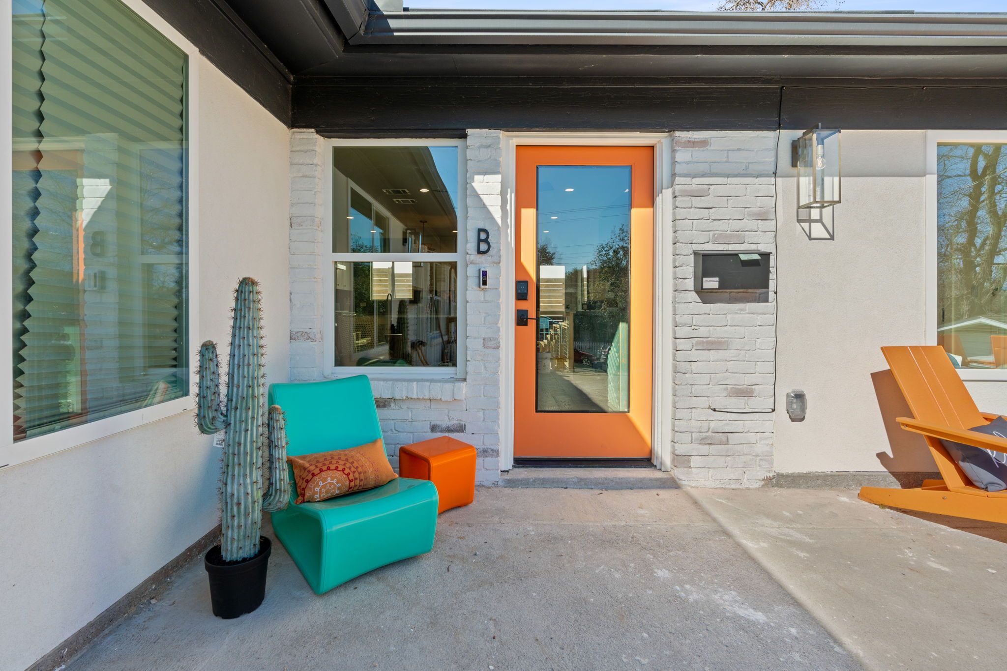 Modern home entrance with bright orange front door, turquoise patio chair, potted cactus, white brick facade and orange Adirondack on a concrete porch.