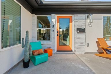 Modern home entrance with bright orange front door, turquoise patio chair, potted cactus, white brick facade and orange Adirondack on a concrete porch.