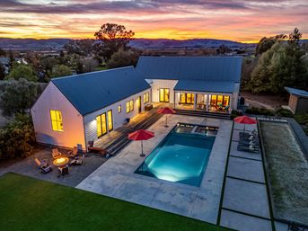 Aerial sunset view of a modern farmhouse with barn-style buildings, warm interior lights, a long illuminated rectangular pool flanked by red umbrellas and lounge chairs, a cozy fire pit seating area, and rolling hills in the distance.