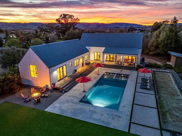 Aerial sunset view of a modern farmhouse with barn-style buildings, warm interior lights, a long illuminated rectangular pool flanked by red umbrellas and lounge chairs, a cozy fire pit seating area, and rolling hills in the distance.