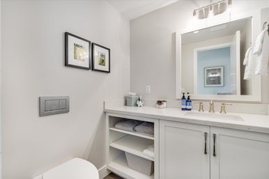 Bright spa-like modern white bathroom with marble vanity, brass faucet, large mirror, open shelves with folded towels and storage basket, and framed wall art