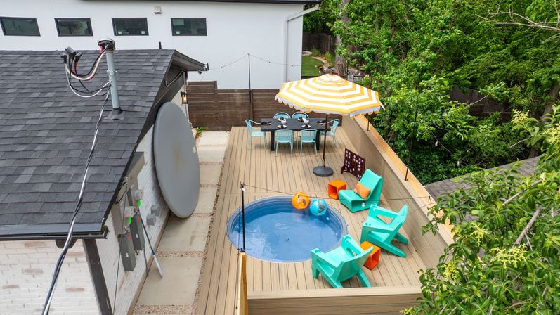 Cheerful residential rooftop deck with a small round plunge pool, turquoise lounge chairs and orange accents, a yellow-striped patio umbrella shading an outdoor dining table, string lights and surrounding trees.