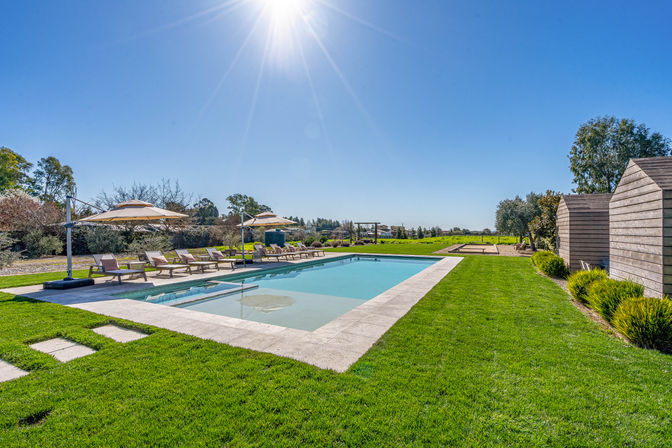 Sun-drenched outdoor rectangular pool with loungers and umbrellas on a manicured green lawn, wooden cabanas at right and open countryside under a clear blue sky.