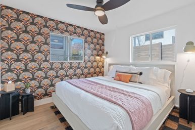 Sunlit modern urban bedroom with white bed and pink quilt, art-deco fan-pattern accent wallpaper, ceiling fan and black bedside tables
