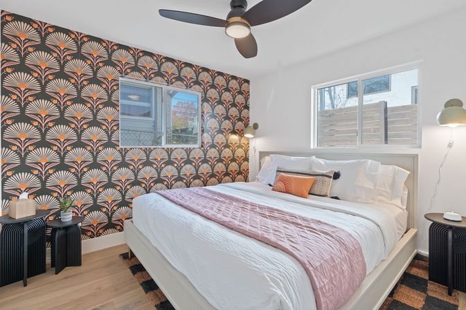 Sunlit modern urban bedroom with white bed and pink quilt, art-deco fan-pattern accent wallpaper, ceiling fan and black bedside tables