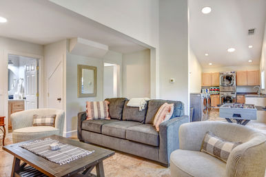 Bright open-plan family room with charcoal sofa, striped throw pillows, two round armchairs and wooden coffee table, opening to a laundry nook with stacked washer-dryer and light wood cabinets.