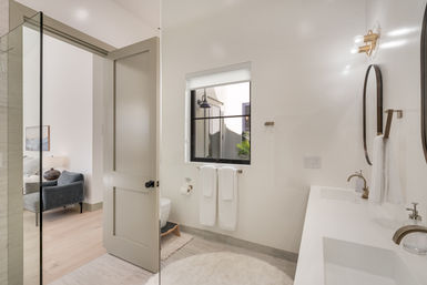 Spa-like modern bathroom with double white vanity and brass faucets, round mirrors, glass shower, white towels and an open door leading to a cozy bedroom.