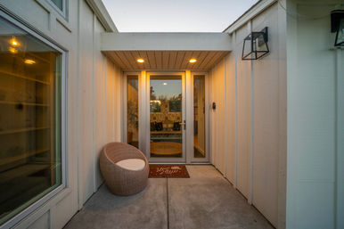 Inviting modern home entry at dusk with glass double doors and sidelights, warm recessed lighting, white board-and-batten siding, a round wicker porch chair on a concrete stoop and wall lanterns casting a cozy glow.
