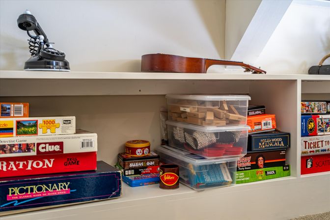 Playroom shelf filled with stacked board game boxes and puzzles, clear plastic storage bins of wooden blocks and game pieces, with a ukulele and vintage rotary-style phone on top.