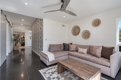 Modern open-plan living room with mauve sectional sofa, wooden coffee table on patterned rug, woven wall baskets, textured accent wall, dark polished concrete floors and ceiling fan