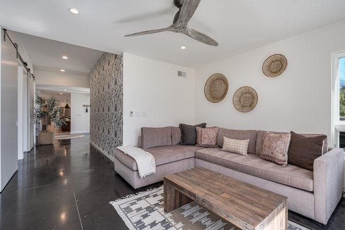 Modern open-plan living room with mauve sectional sofa, wooden coffee table on patterned rug, woven wall baskets, textured accent wall, dark polished concrete floors and ceiling fan