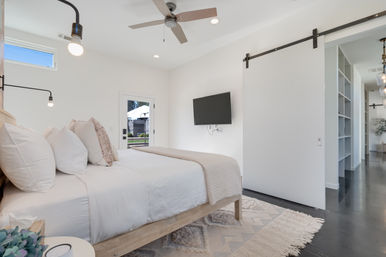 Sunlit modern bedroom interior with white bedding on a wooden platform bed, ceiling fan, wall-mounted TV, sliding barn door to hallway, neutral rug and pendant lights.