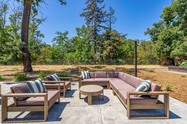 Sunlit backyard concrete patio with modern wooden L-shaped sectional, matching armchairs, round coffee table and striped cushions surrounded by mature trees