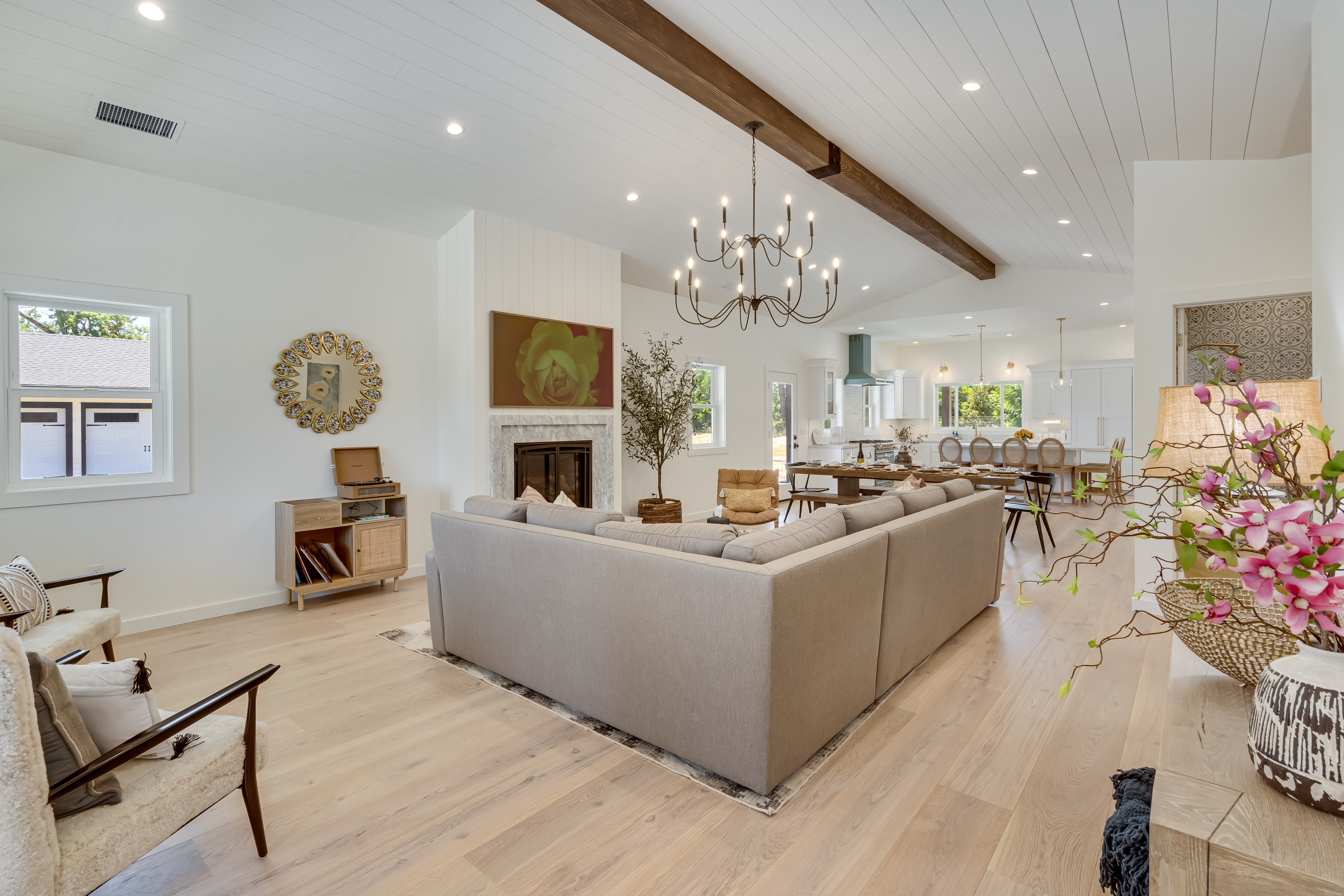 Sunlit open-concept modern farmhouse living room and kitchen with a large gray sectional, vaulted white shiplap ceiling with exposed dark wood beam and chandelier, marble fireplace, light hardwood floors, and dining area in the background.