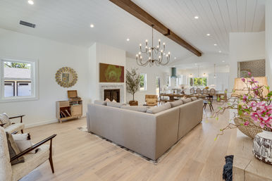 Sunlit open-concept modern farmhouse living room and kitchen with a large gray sectional, vaulted white shiplap ceiling with exposed dark wood beam and chandelier, marble fireplace, light hardwood floors, and dining area in the background.