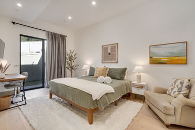 Modern sunlit bedroom with green quilted bed and rolled white towels, bedside tables with lamps, sliding glass door and gray curtains to a patio, desk, potted plant, comfy armchair, and abstract wall art.