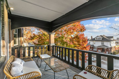 Covered second-floor porch with arched openings, rattan seating, cushions and glass-top table overlooking a suburban street and vibrant orange-red fall foliage near brick houses