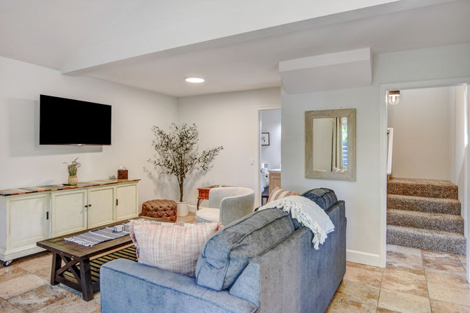 Cozy basement living room with a gray sofa and throw, wall-mounted flat-screen TV above a cream media console, rustic coffee table, armchair and tufted ottoman, potted tree, wall mirror, and carpeted stairs.