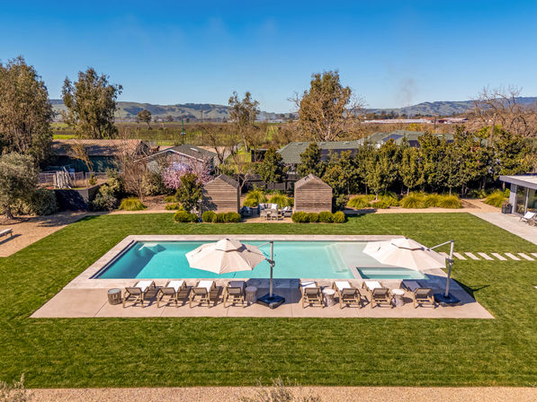 Aerial view of a modern rectangular outdoor pool with sun-ready loungers and large umbrellas on a concrete deck, surrounded by a manicured lawn, small garden huts and rolling hills in the distance.