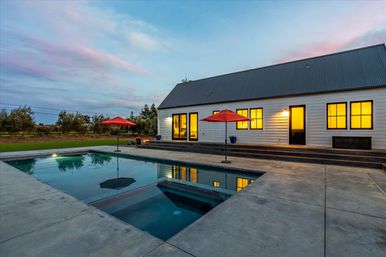 Modern farmhouse backyard at dusk with warm lit windows, rectangular swimming pool reflecting a pink-blue sky, concrete patio and red patio umbrellas.