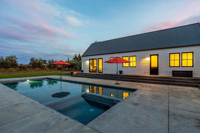 Modern farmhouse backyard at dusk with warm lit windows, rectangular swimming pool reflecting a pink-blue sky, concrete patio and red patio umbrellas.