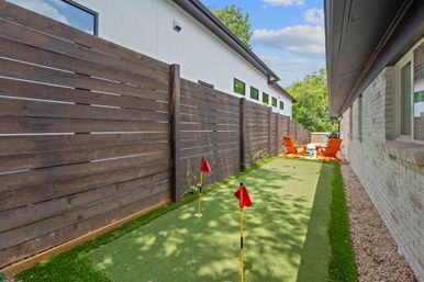 Narrow backyard with artificial putting green between a white brick house and tall dark wood fence, three red-flag golf holes and two bright orange lounge chairs at the far end under a blue sky.