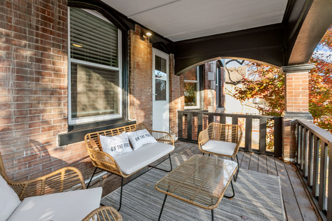 Inviting covered brick front porch with rattan sofa and chairs, glass-top coffee table, striped rug and autumn foliage