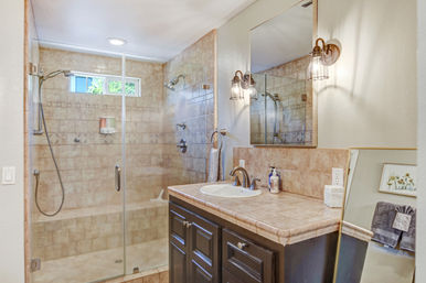 Bright home bathroom with glass-enclosed tiled walk-in shower featuring dual showerheads and bench, next to a beige-tiled vanity with sink, mirror, bronze fixtures and wall sconces.