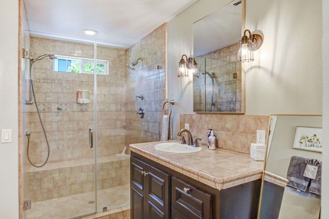 Bright home bathroom with glass-enclosed tiled walk-in shower featuring dual showerheads and bench, next to a beige-tiled vanity with sink, mirror, bronze fixtures and wall sconces.