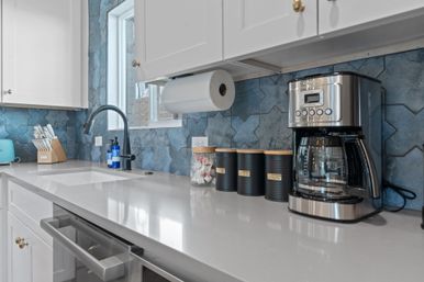 Modern kitchen countertop with white cabinets and quartz surface, blue geometric tile backsplash, undermount sink with matte-black faucet, stainless-steel coffee maker, black labeled canisters and knife block.