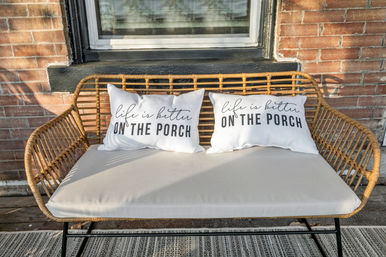 Cozy wicker porch bench with beige cushion and two white throw pillows that read 'Life is better on the porch,' set against a brick wall beneath a window.