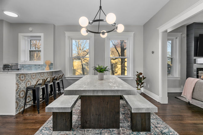 Bright modern open-concept dining room featuring a concrete tabletop with matching benches, round globe chandelier, large double windows, patterned rug and hardwood floors, plus an adjacent tiled breakfast bar with metal stools.