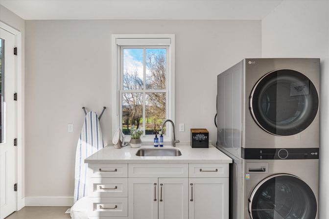 Sunny modern laundry room with stacked stainless washer and dryer, white cabinets and sink beneath a window overlooking trees, striped ironing board.