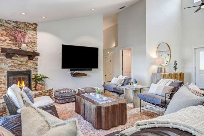 Cozy modern living room with stone fireplace and wall-mounted TV, live-edge wood coffee table, neutral sofas and accent chairs on a patterned rug with plants and decorative accents.