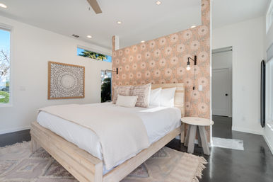 Sunlit modern bedroom with a light-wood platform bed and crisp white linens against a peach floral wallpaper accent wall, black wall sconces, round wooden side table, large windows and a textured rug.