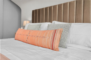 Modern cozy bedroom interior with a tall taupe velvet channel headboard, crisp white bedding, textured gray pillows and a long coral-orange lumbar pillow, bedside lamp and arched wall niche.