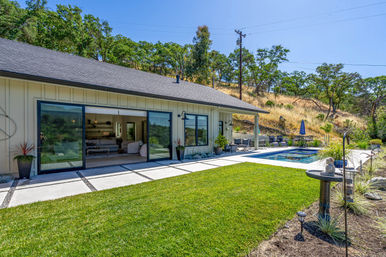 Sunny backyard oasis of a modern single-story home with open sliding glass doors, manicured lawn and concrete pavers, rectangular pool with lounge chairs and umbrella, set against an oak‑dotted hillside