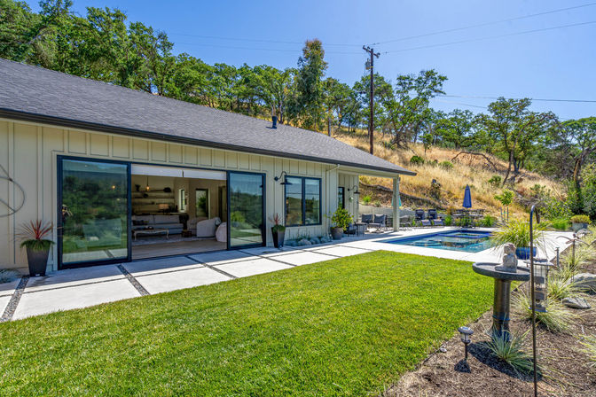 Sunny backyard oasis of a modern single-story home with open sliding glass doors, manicured lawn and concrete pavers, rectangular pool with lounge chairs and umbrella, set against an oak‑dotted hillside