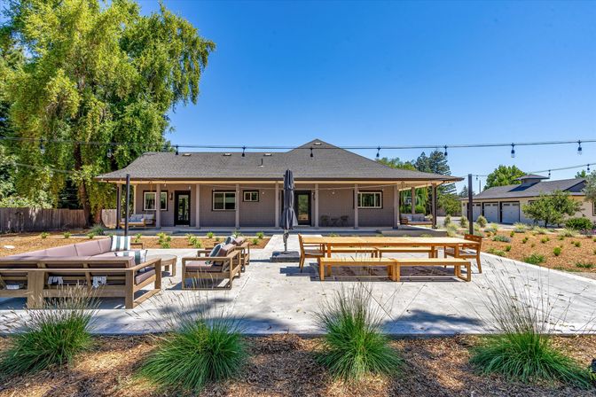 Spacious suburban backyard patio with single-story house and covered porch, concrete patio with string lights, wooden picnic table and cushioned lounge seating, landscaped yard and mature trees under a clear blue sky