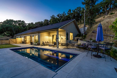 Dusk view of a modern hillside ranch-style home with a cozy lit patio, outdoor dining set and a reflecting rectangular pool.