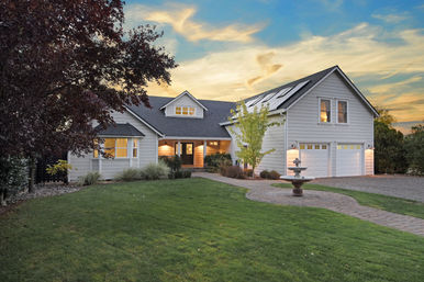 Charming suburban home with white siding and rooftop solar panels, lit windows, attached two-car garage, manicured lawn, paver walkway and fountain at sunset.