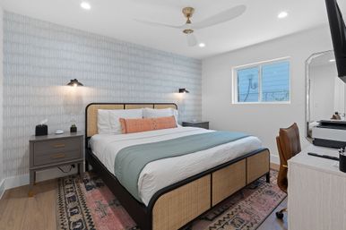 Bright modern coastal-style bedroom with rattan king bed dressed in white linens, green throw and coral lumbar pillow, patterned area rug, matching nightstands with wall sconces, ceiling fan, small window, and a desk with leather chair and wall-mounted TV.