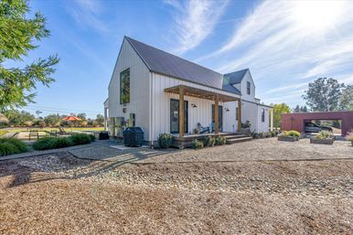 Cheerful modern white farmhouse with metal roof and covered wooden porch strung with lights, gravel courtyard with raised planters, nearby outdoor seating and carport under a bright blue sky.