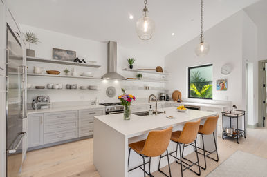 Bright modern open-concept kitchen with white island and sink, stainless range hood, leather bar stools, glass pendant lights, open shelving, light wood floors, and a vase of colorful flowers
