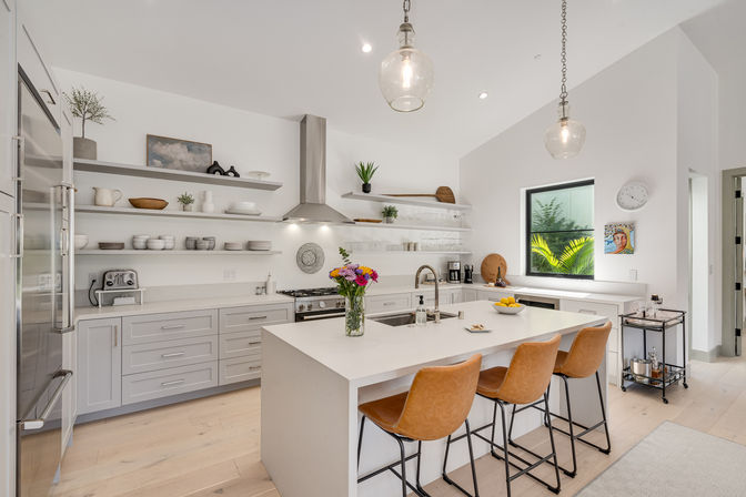 Bright modern open-concept kitchen with white island and sink, stainless range hood, leather bar stools, glass pendant lights, open shelving, light wood floors, and a vase of colorful flowers