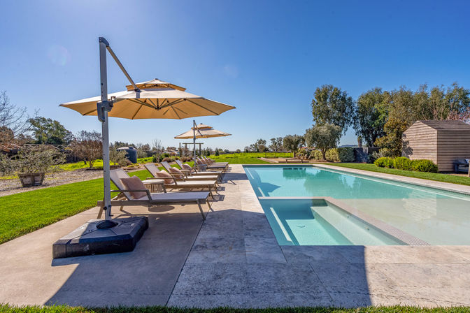 Sunny outdoor rectangular swimming pool with built-in steps and clear water, lined with beige lounge chairs and large cantilever umbrellas on a stone patio, surrounded by manicured lawn, trees and open countryside under a bright blue sky.